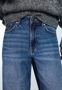 Kék magas derekú farmernadrág egyenes szárakkal, finom kopásokkal és rézszínű varrással. Szürke kötött pulóverrel viselve.