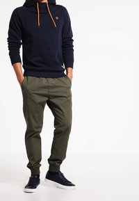 Navy hoodie med orange dragkedjor och diagonala sömmar, tillsammans med olivgröna tapered-byxor och mörka sneakers. Minimalistisk design.