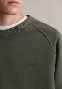 Oliwkowa bluza z textured materiału, ze ściągaczem przy kołnierzu i rękawami raglanowymi. Posiada subtelne logo wytłoczone na piersi.