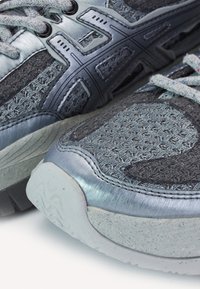 Gros plan sur des baskets athlétiques grises et argentées avec tissu tricoté, surpiqûres synthétiques et semelles blanches texturées sur une surface blanche.