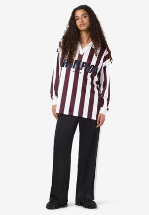 Vrouw met lang krullend haar, gekleed in een langermige bordeauxrode en witte gestreepte blouse met de tekst "CHAMPION" en zwarte wijde broek.