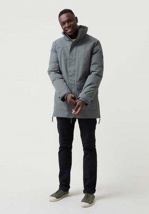 Parka gris, acolchada, con un alto cuello, cremallera frontal y bolsillos ocultos, combinada con pantalones negros y zapatos verdes.