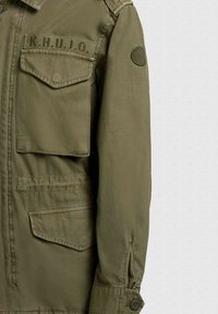 Vojaški jakna olivno zelene barve s preklopnimi žepi, gumbnimi manšetami, okroglim obližem na rokavu in vezenim napisom "K.H.U.J.O." nad prsnim žepom.