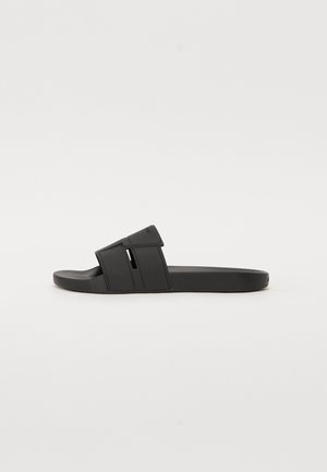 Sandale mule noire avec une large bride comportant des découpes et une semelle intérieure texturée sur une semelle plate.