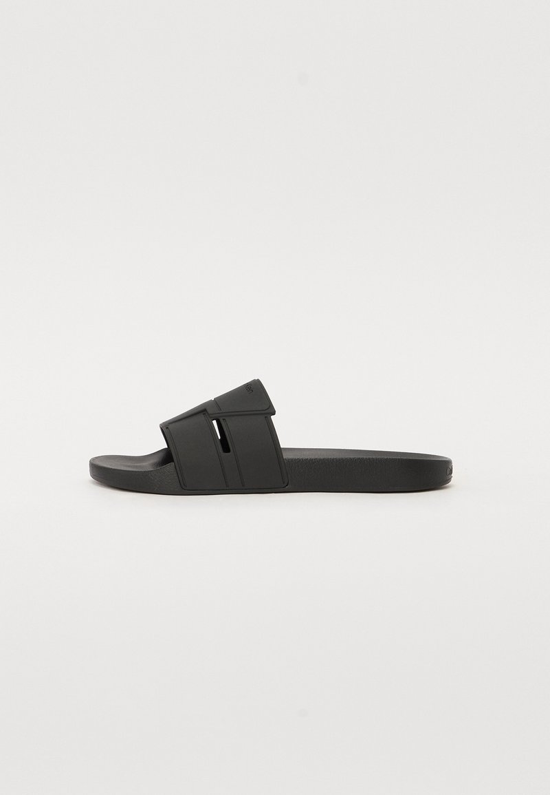 Sandale mule noire avec une large bride comportant des découpes et une semelle intérieure texturée sur une semelle plate.