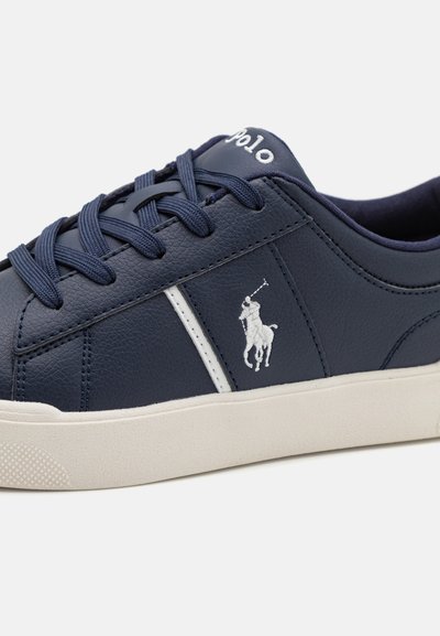Zapatillas de cuero azul marino con franja lateral blanca, punta redonda y suela de goma. Presenta un logo de polo bordado en el lateral y diseño de cordones.