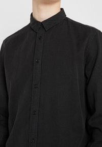Czarna koszula zapinana na guziki z klasycznym kołnierzykiem, o gładkiej fakturze i subtelnym połysku, podkreślona czarnymi guzikami z przodu.