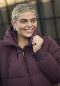 Bordový puffrový kabát s vysokým golierom, zipsom a ribbovanými manžetami. Má hladkú textúru a kapucňu. Model sa usmieva.