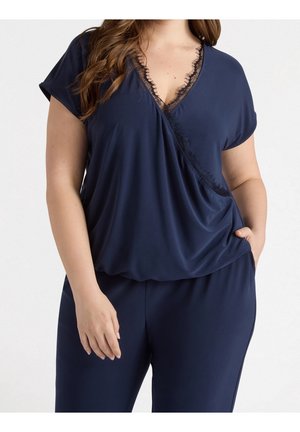 Femme portant un chemisier bleu marine à manches courtes avec un col en V orné de dentelle et un pantalon assorti, main dans la poche, sur fond blanc uni.