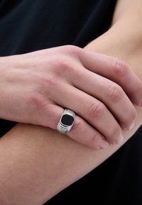Sidabrinis žiedas su tekstūruotu juostele ir kvadratiniu juodu akmeniu, įstatytu centre, dėvimas ant rankos prieš šviesią odą.