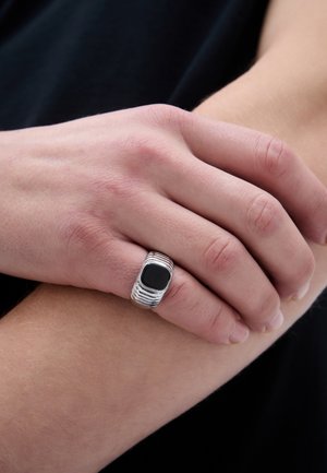 Stříbrný prsten s texturovaným páskem a čtvercovým černým kamenem zasazeným ve středu, nošený na ruce s světlou pletí.