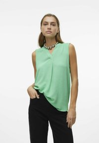 Haut sans manches vert clair avec un col en V, fabriqué en tissu texturé. Associé à un pantalon noir, avec un collier en perles métalliques rondes.