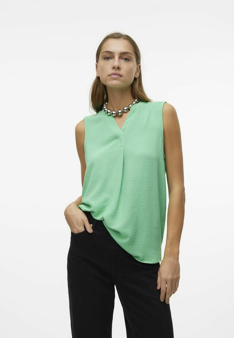 Haut sans manches vert clair avec un col en V, fabriqué en tissu texturé. Associé à un pantalon noir, avec un collier en perles métalliques rondes.
