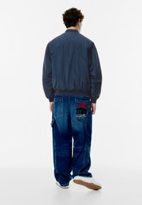 Marine bomberjack, donkere denim jeans met grafisch zakontwerp, en witte sneakers. Glad materiaal, relaxte pasvorm en elastische manchetten.