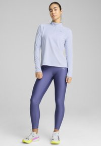 Haut à manches longues violet clair avec un col zippé, associé à des leggings moulants violet foncé. Les chaussures sont blanches avec des accents jaunes et roses.