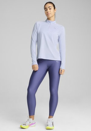 Haut à manches longues violet clair avec un col zippé, associé à des leggings moulants violet foncé. Les chaussures sont blanches avec des accents jaunes et roses.
