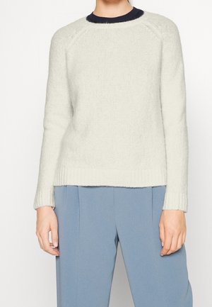 Persoon draagt een witte gebreide trui met geribde boorden en zoom, gecombineerd met lichtblauwe hoge taille broek tegen een effen achtergrond.