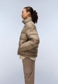 Beige pufferjack met hoge kraag, voorzien van een ritsluiting en zijzakken, gedragen over een gestreept shirt. Glad materiaal, gewatteerd ontwerp.