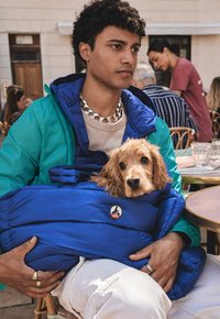 Borsetta per animali domestici imbottita blu con un logo circolare, tenuta da una persona che indossa una giacca verde con interno blu. Un cocker spaniel sbircia fuori.