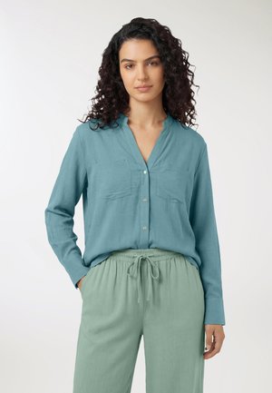 Donna con capelli ricci scuri che indossa una camicetta verde acqua con bottoni e pantaloni verdi chiaro con coulisse, in piedi con una mano in tasca su sfondo bianco.