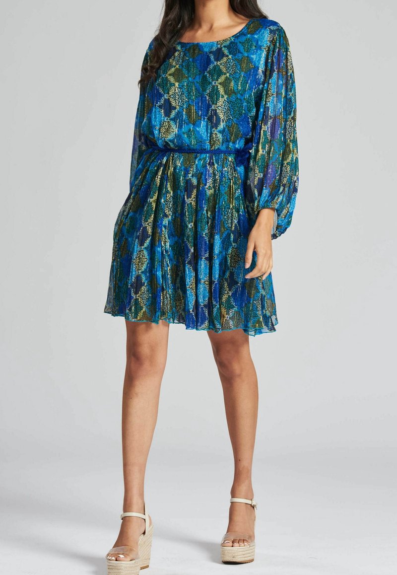 Femme portant une robe bleu et vert à motifs, longueur genou, avec manches longues et sandales compensées beiges.
