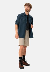 Camicia a maniche corte blu navy con due tasche sul petto, abbinata a pantaloni corti beige chiaro e scarponi da escursionismo marroni con calze nere a fantasia.