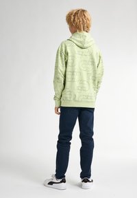 Sudadera verde con capucha, adornada con patrones geométricos negros; combinada con jeans azul oscuro y zapatillas blancas con toques negros.