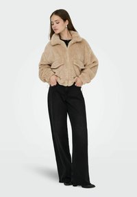 Veste zippée en fausse fourrure beige avec un col, deux poches avant et une coupe courte, associée à un jean noir taille haute à jambes larges.