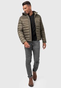 Chaqueta acolchada verde oliva con costuras horizontales, camiseta negra de cuello en V, pantalones gris ajustados con detalles acanalados y botas marrón de tobillo.