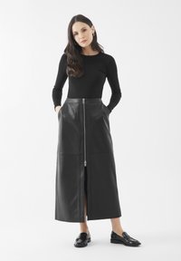 Vrouw met lang, donker haar in een zwart lange mouw top, zwarte leren midi rok met rits aan de voorkant en zwarte loafers, staat met haar handen in de zakken.