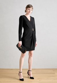 Robe noire portefeuille à manches longues, ceinturée à la taille avec une boucle dorée. Associée à une pochette noire et des escarpins noirs à talons aiguilles.