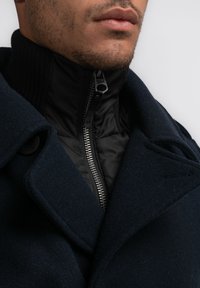 Abrigo de lana Navy con cuello alto, interior acolchado negro y una cremallera plateada brillante con un tirador de marca.