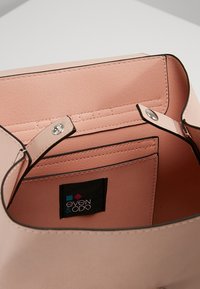 Vaaleanpunainen käsilaukku, jossa on sileä pinta, kaksi nepparikiinnitystä, vetoketjutasku sisäpuolella ja merkkilaatta. Reunoissa on kontrastivärinen musta reunus.