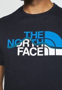 Camiseta de manga corta azul marino oscuro de algodón con un gran logo impreso en blanco y azul que presenta un diseño ondulado a lo largo del texto.