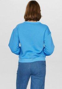 Mėlynas džemperis su apvalia iškirpte, raglan rankovėmis ir banguota apykakle. Minkšta medžiaga su lygiu paviršiumi. Dėvimas su mėlynomis džinsinėmis kelnėmis.