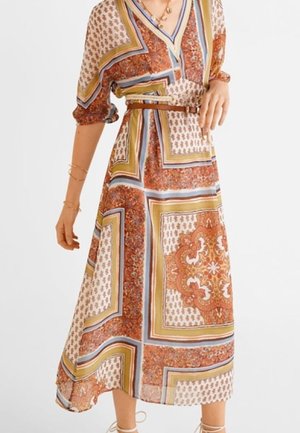 Mujer con vestido midi estampado con diseños geométricos y florales en tonos marrón, beige y azul, con cinturón en la cintura, de pie con una mano en la cadera.