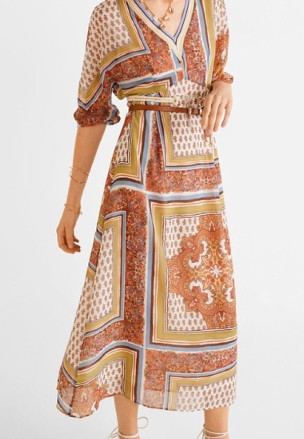 Femme portant une robe midi à motifs avec des dessins géométriques et floraux marron, beige et bleu, ceinturée à la taille, debout avec une main sur la hanche.