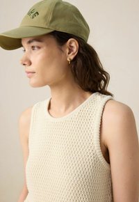 Beige knitted sleeveless top with a textured pattern, paired with a light green cap featuring embroidered text. Gold hoop earrings accent the look.