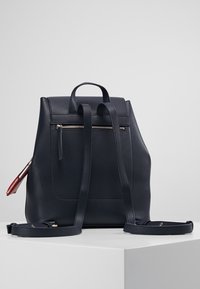 Zaino in pelle blu navy con texture liscia, tasca verticale con zip, spallacci regolabili e silhouette arrotondata. Dettaglio della zip rossa sul lato.