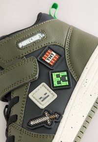 Scarpa da ginnastica verde e nera con cinturino in velcro, caratterizzata da patch di gomma testurizzati: TNT, un volto, un blocco e una spada. Suola bianca con macchie.