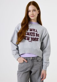 Sudadera gris corta con el texto "SIEMPRE SERÁ NUEVA YORK" en color burdeos, superpuesta a una blusa lila; combinada con jeans grises.
