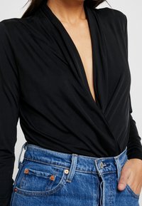 Schwarzes Langarm-Wickeloberteil mit tiefem V-Ausschnitt; glattes Material; kombiniert mit hochgeschnittenen blauen Jeans aus Denim mit bronzefarbenen Hardware und Nähten.