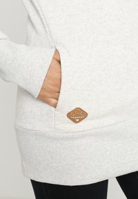 Vaaleanharmaa huppari, jossa on ribbireunukset hihoissa ja helmassa. Hupparissa on sivutasku ja ruskea brändipatch, jossa on timantin muoto ja teksti.