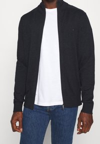 Giacca zip grigia scura con colletto alto, polsini a costine e un logo discreto. Indossata sopra una maglietta bianca e abbinata a jeans blu.