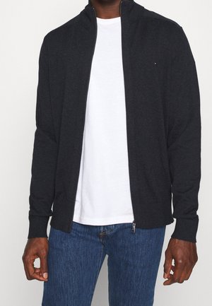 Giacca zip grigia scura con colletto alto, polsini a costine e un logo discreto. Indossata sopra una maglietta bianca e abbinata a jeans blu.
