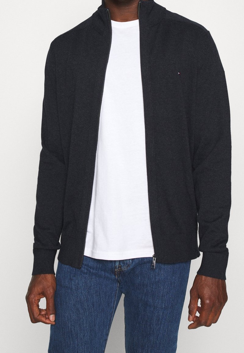 Giacca zip grigia scura con colletto alto, polsini a costine e un logo discreto. Indossata sopra una maglietta bianca e abbinata a jeans blu.