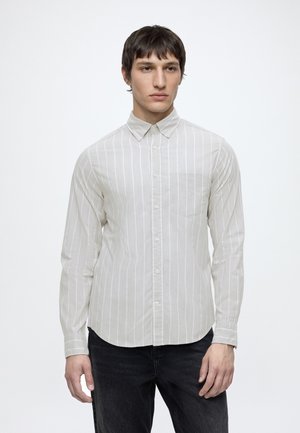 Homme aux cheveux courts et foncés portant une chemise blanche à manches longues avec de fines rayures verticales et un pantalon foncé, debout devant un fond uni.