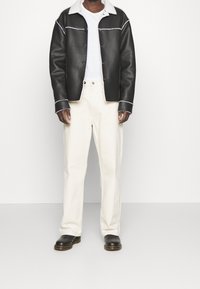 Persona che indossa una giacca di pelle nera con cuciture bianche, una camicia bianca, pantaloni bianco sporco e scarpe di pelle nere, in piedi su uno sfondo neutro.