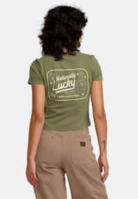 Oliwkowa, krótkotidowa koszulka z białym grafiką tekstu "Naturally Lucky Club" i symbolami. Posiada krótkie rękawy i okrągły dekolt.