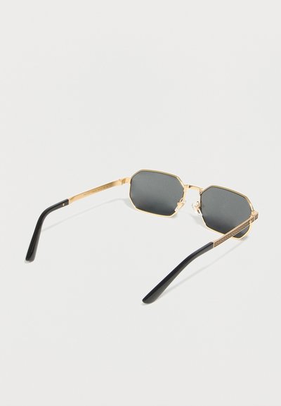 Οκταγωνικά γυαλιά ηλίου με χρυσή κορνίζα, σκούρους φακούς και μαύρους βραχίονες, εκτεθειμένα σε λευκό φόντο.